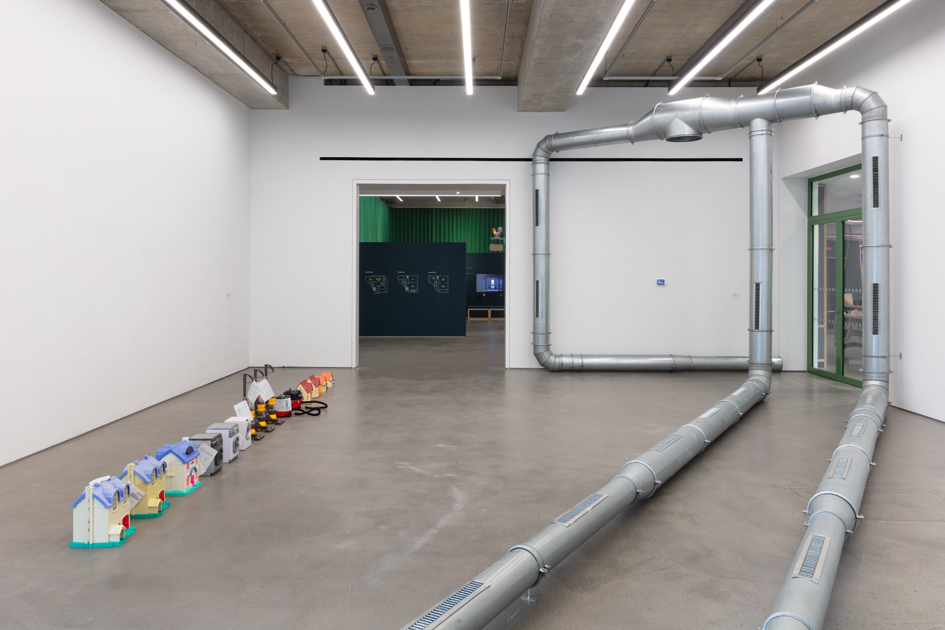 A straight line of plastic children's toys including dolls houses and replicas of vacuum cleaners and washing machines. Large metal piping from an air condition unit is also assembled across the room.